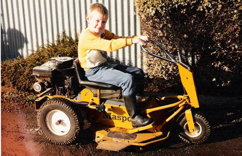 Mike as a boy on a yellow Masport ride-on mower, gumboots on the footplate